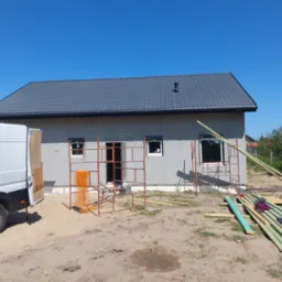 Budynek jednorodzinny w trakcie budowy z szarym tynkiem, czarną dachówką i rusztowaniami, widoczny biały van z otwartymi drzwiami i stertą drewna na pierwszym planie.