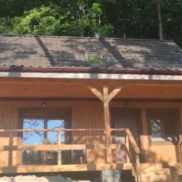 Drewniany domek letniskowy z ciemną dachówką, widoczny z zewnątrz, z przyczepą kempingową Fendt w tle po lewej stronie, otoczony drzewami.