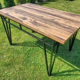 Stół z blatem z ciemnego drewna i czarnymi metalowymi nogami, ustawiony na trawniku w ogrodzie.