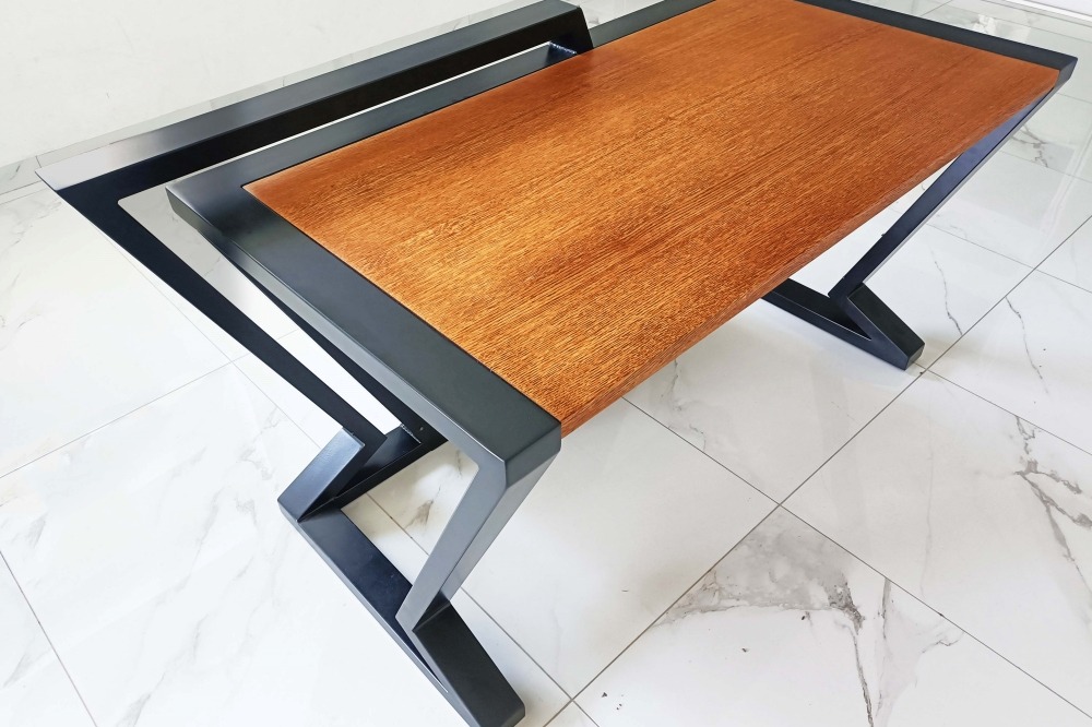 Stolik kawowy z blatem z drewna o ciepłym odcieniu i czarną, geometryczną podstawą z metalu na tle marmurowej podłogi.