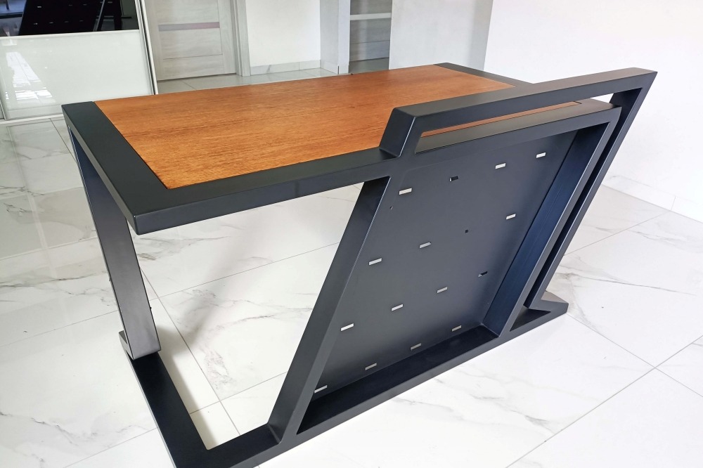 Nowoczesny stół z drewnianym blatem i metalową, geometryczną podstawą w kolorze czarnym, widoczne otwory na śruby w metalowej konstrukcji, biała marmurowa podłoga w tle.