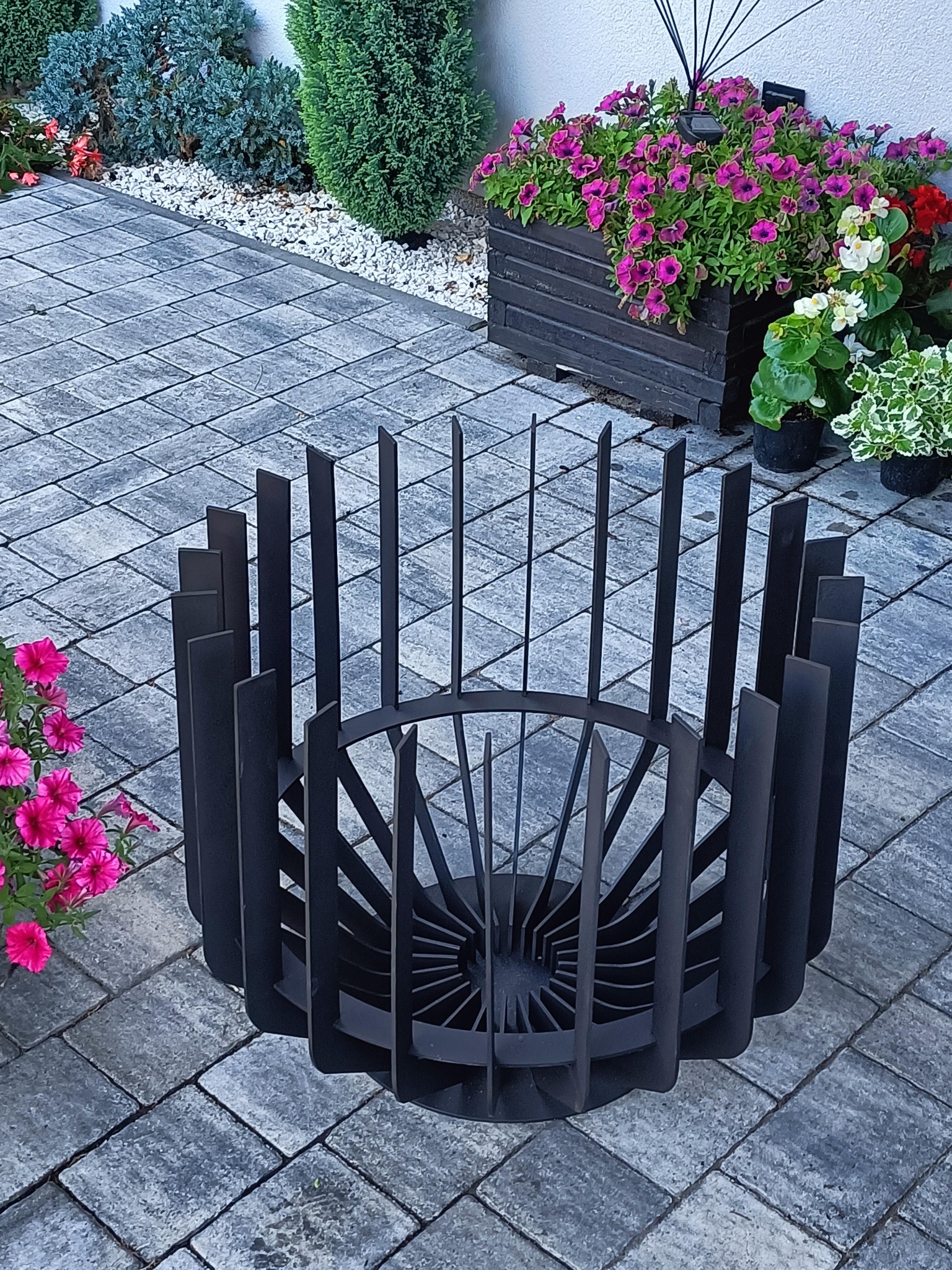 Nowoczesny, metalowy kosz na ognisko, otoczony kwiatami w donicach i na rabatach, ułożony na szarej kostce brukowej.