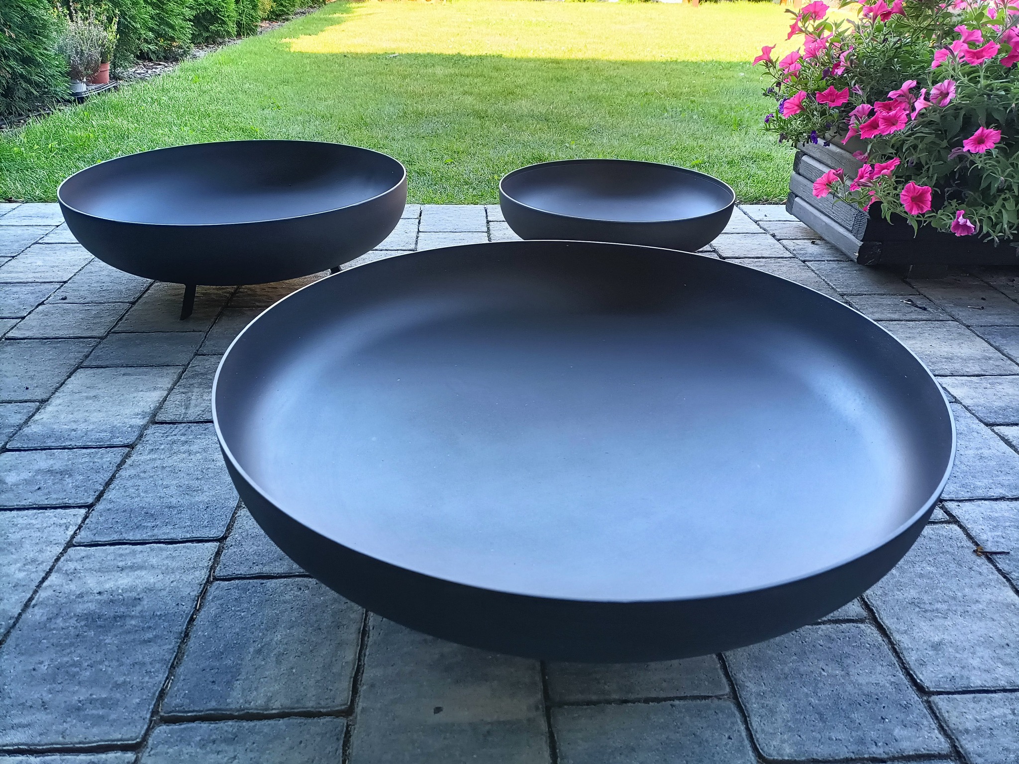 Trzy czarne, metalowe misy ogrodowe o różnych rozmiarach, ustawione na szarej kostce brukowej, z zielonym trawnikiem i krzewami w tle oraz skrzynką z różowymi kwiatami po prawej stronie.
