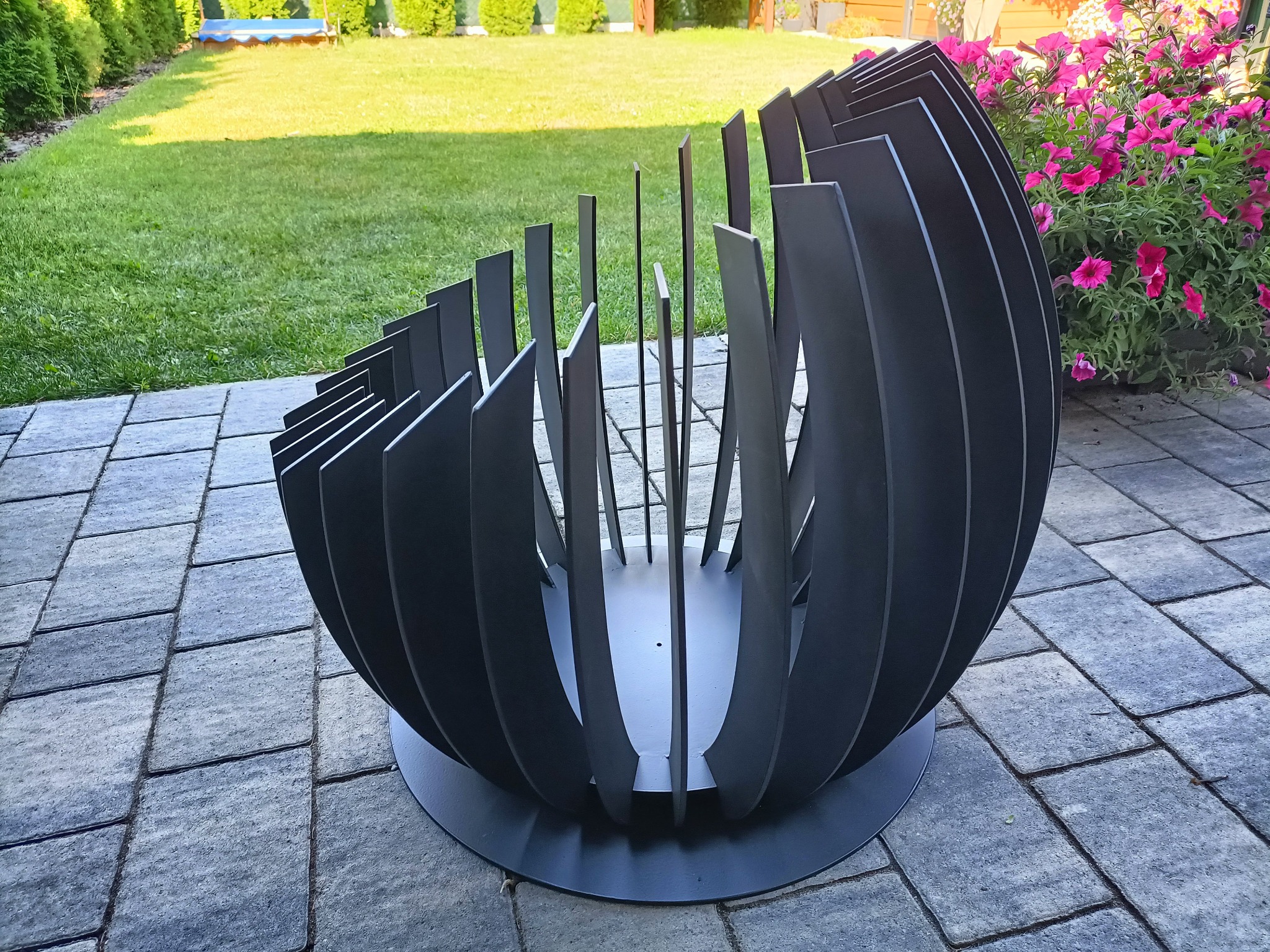 Nowoczesny, metalowy kosz na drewno kominkowe w kształcie półkuli, ustawiony na szarym bruku w ogrodzie z zielonym trawnikiem i kwitnącymi różowymi kwiatami w tle.