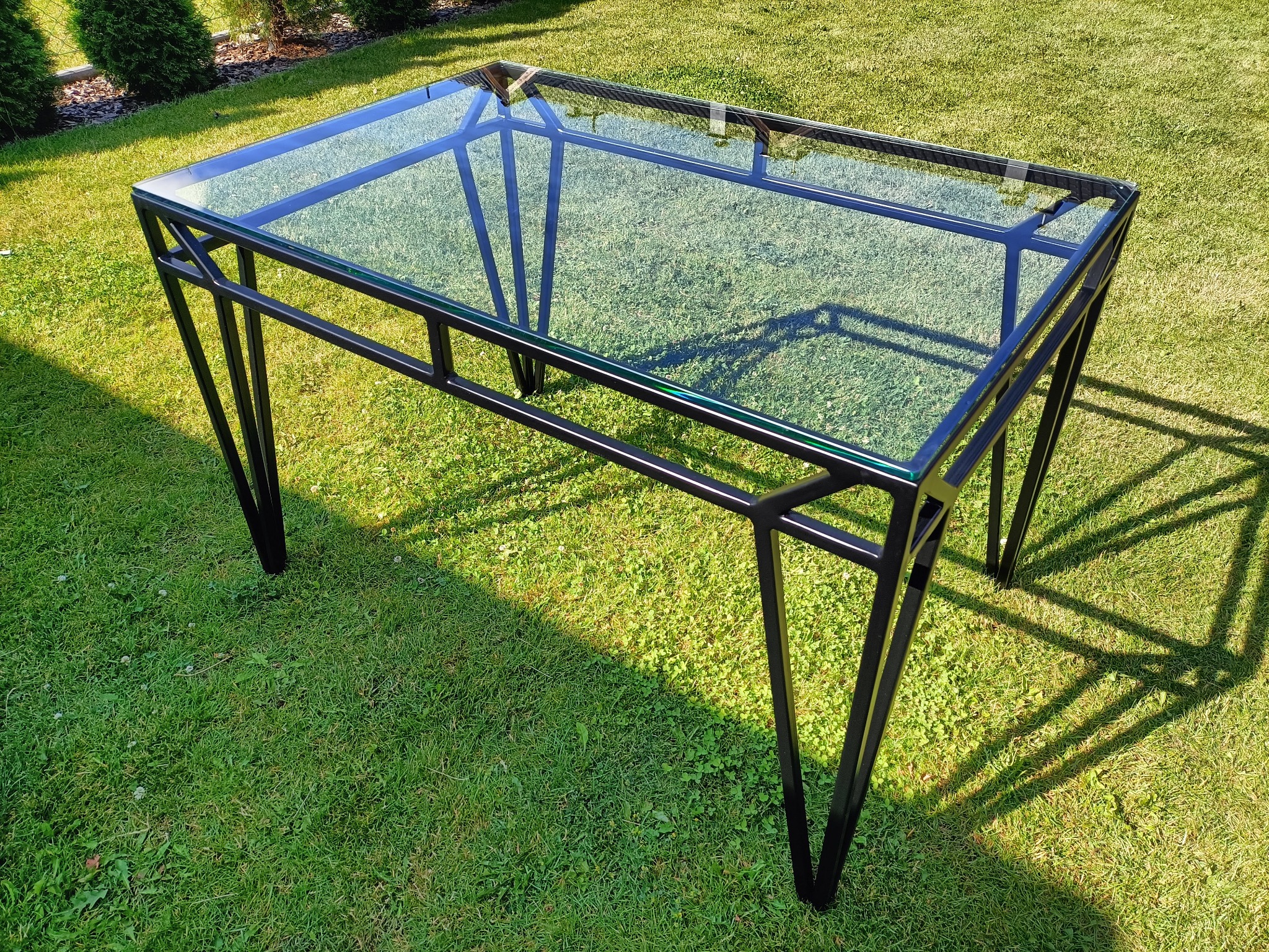 Stół z czarną metalową ramą i szklanym blatem, ustawiony na trawniku w słoneczny dzień, widoczne cienie rzucane przez nogi stołu na zieloną trawę.