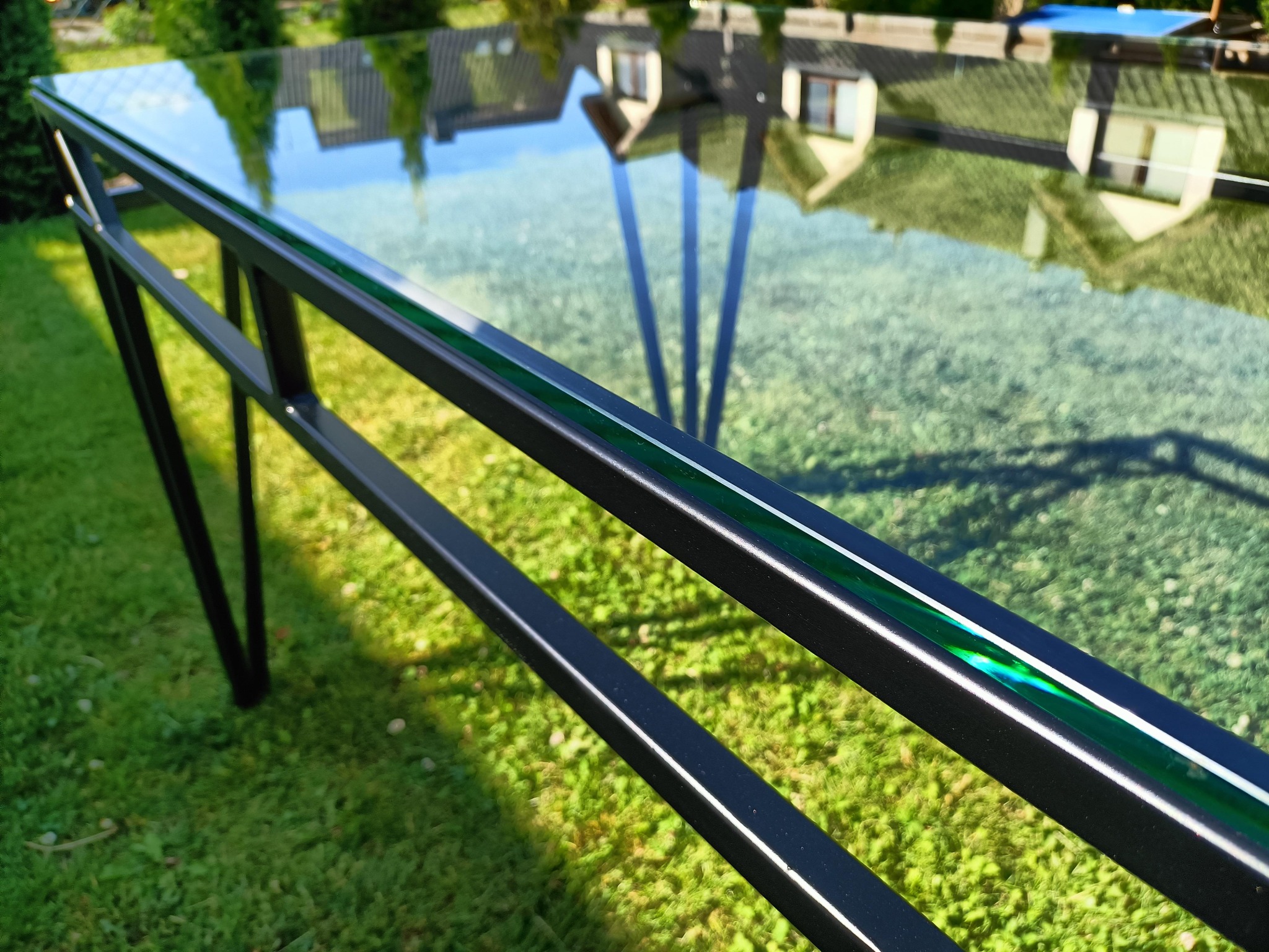 Szklany blat stołu z czarną metalową ramą, odbijający budynki i drzewa na trawniku.