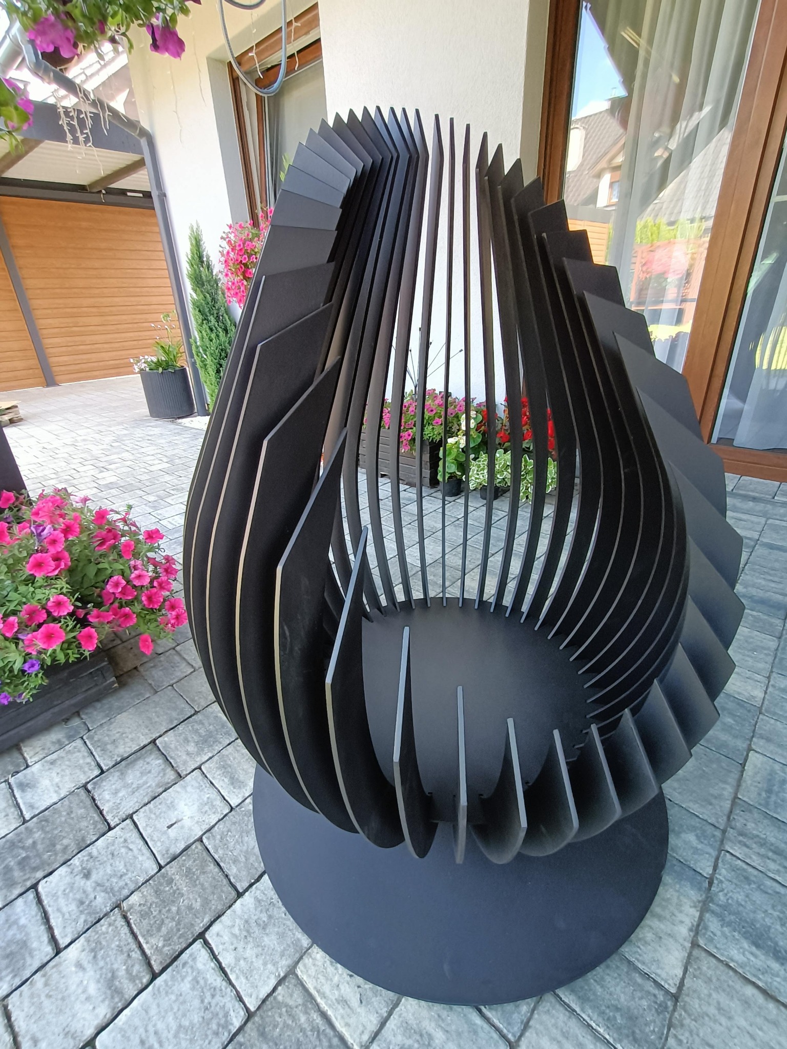 Nowoczesny, ażurowy, metalowy kosz na drewno kominkowe w kształcie łzy, ustawiony na szarym bruku przed domem z drewnianą bramą garażową i oknami z brązowymi ramami. W tle widoczne kwiaty w donicach.