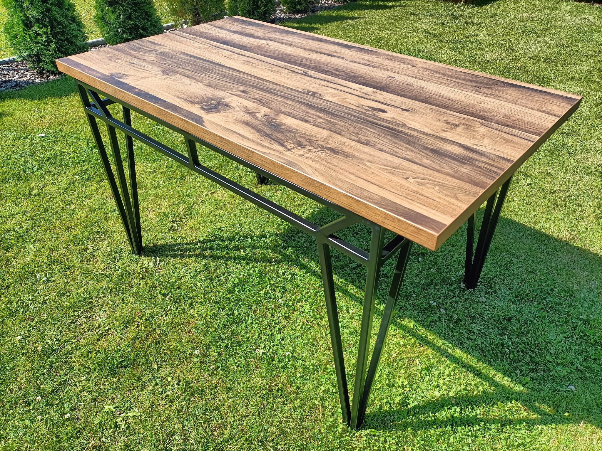 Stół z blatem z ciemnego drewna i czarnymi metalowymi nogami, ustawiony na trawniku w ogrodzie.