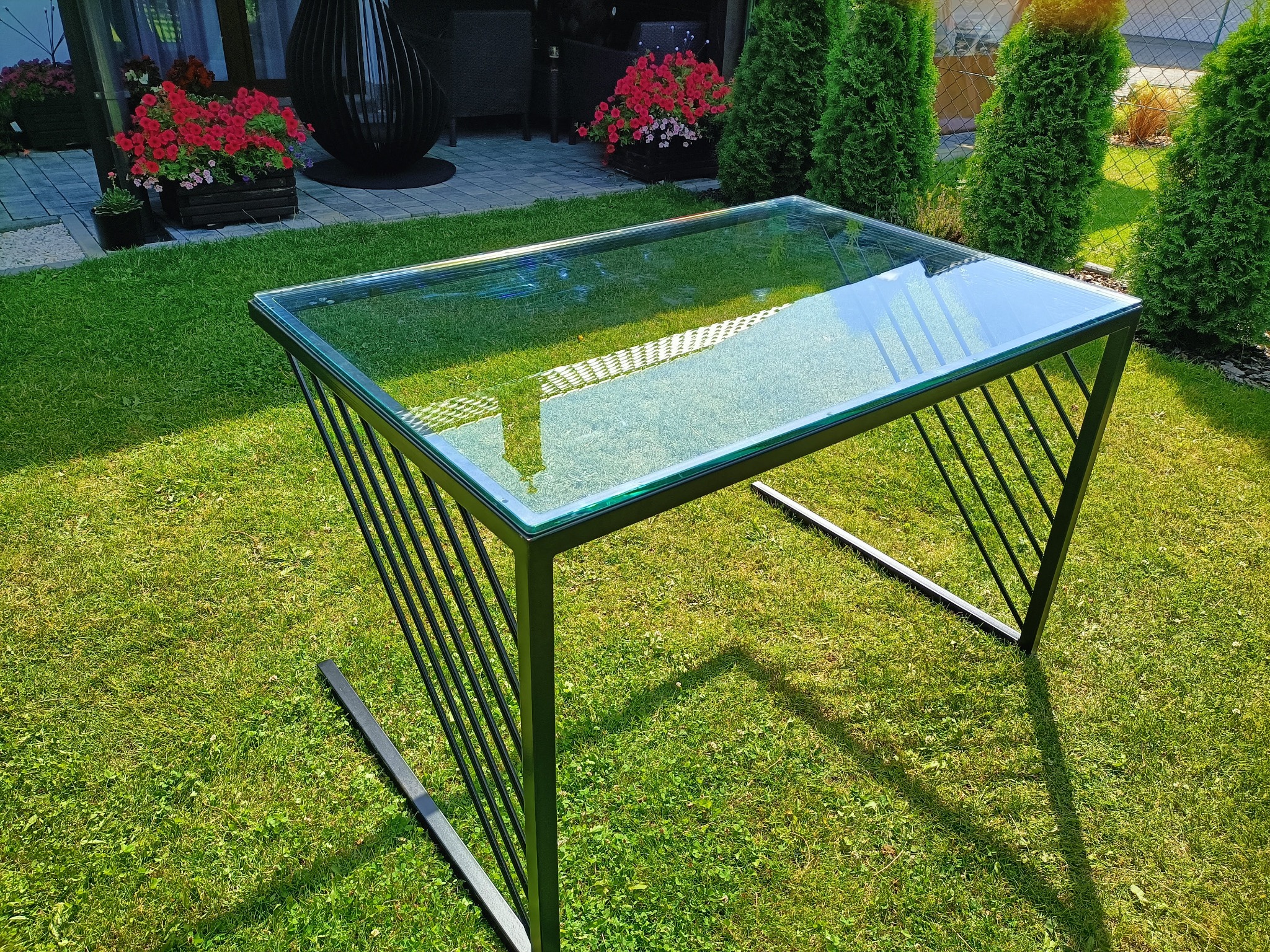 Szklany stolik kawowy z czarną, geometryczną podstawą z metalowych prętów, ustawiony na trawniku w ogrodzie. W tle widoczne tuje i kwiaty.