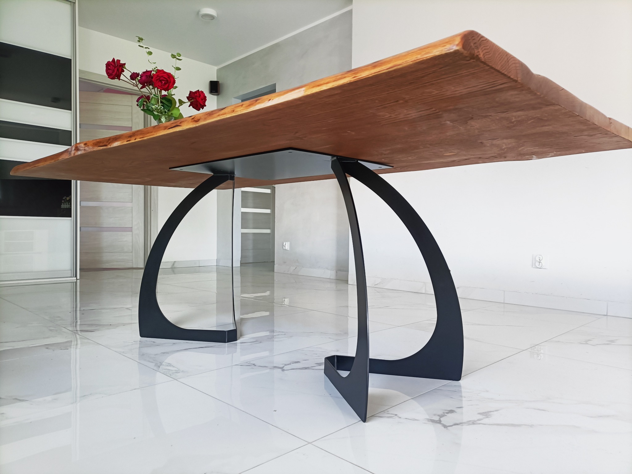 Stół z blatem z litego drewna o nieregularnych krawędziach, wsparty na czarnych, łukowatych nogach, na tle białych płytek podłogowych i bukietu czerwonych róż.