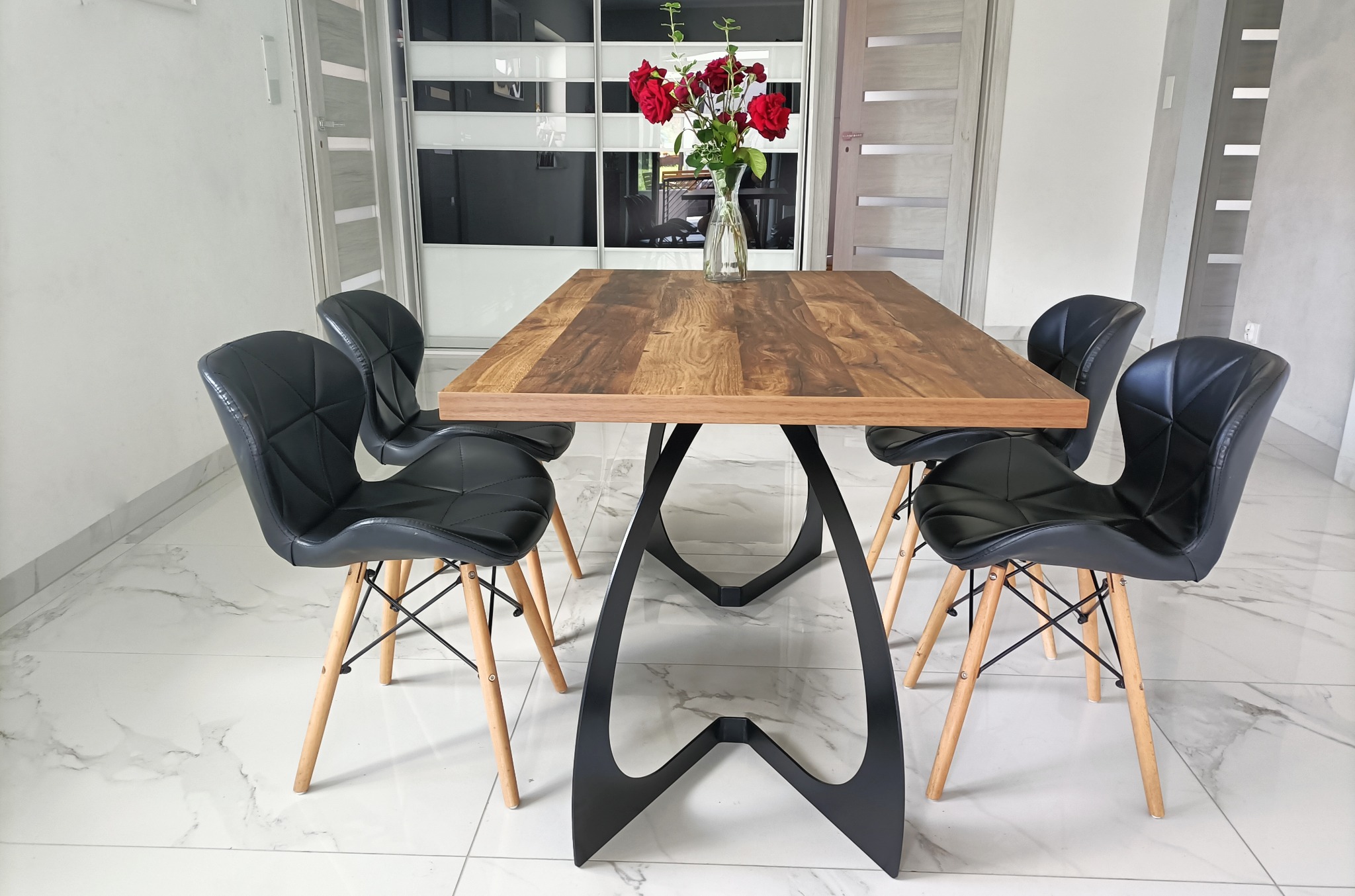 Stół z blatem z drewna i czarną, metalową podstawą, otoczony czterema czarnymi krzesłami z drewnianymi nogami, na marmurowej podłodze, z wazonem czerwonych róż na blacie, na tle białych drzwi i szafy.
