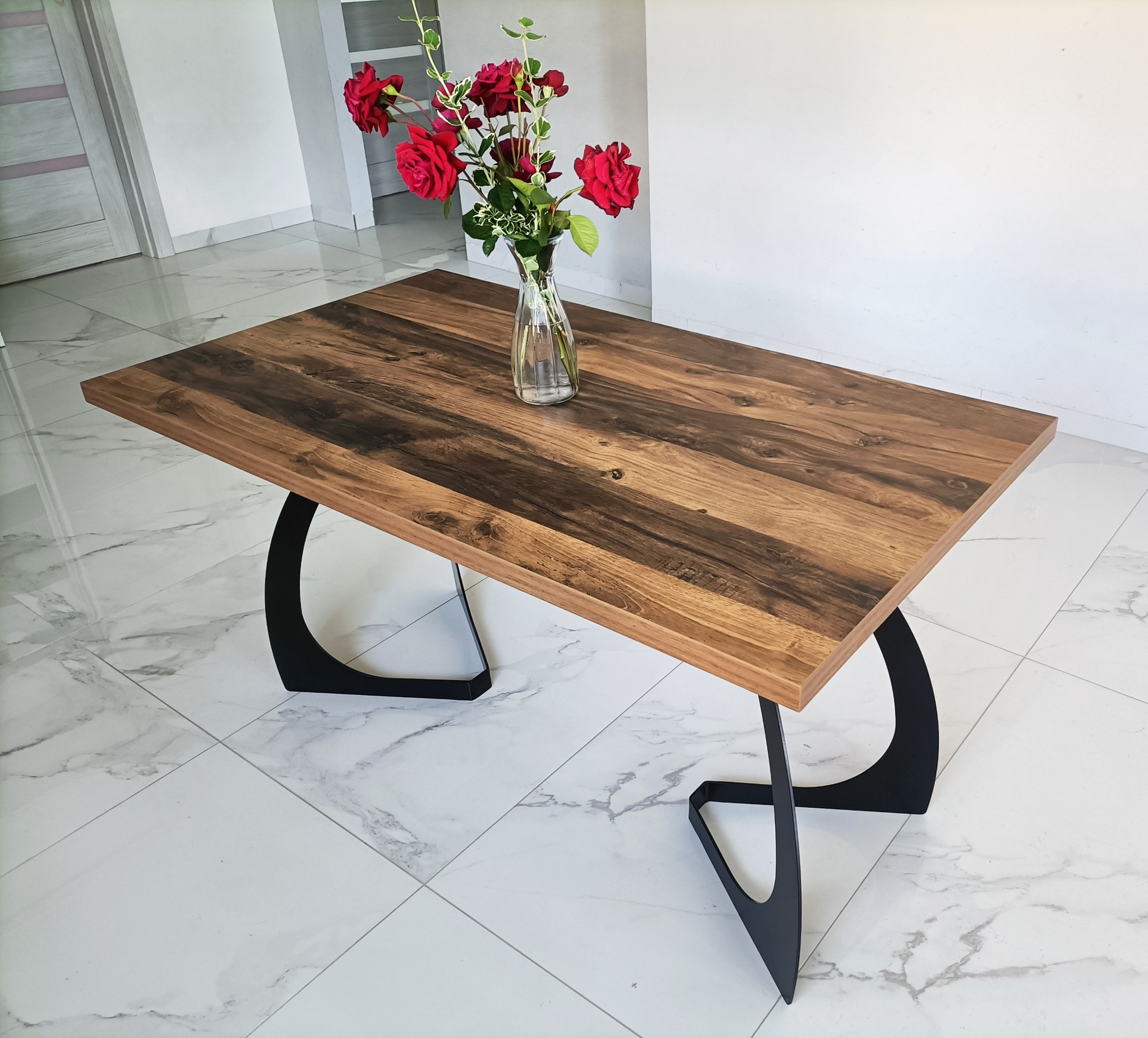 Stolik kawowy z blatem z ciemnego drewna i czarnymi, zakrzywionymi nogami, na podłodze z białych płytek imitujących marmur, z wazonem czerwonych róż na blacie.