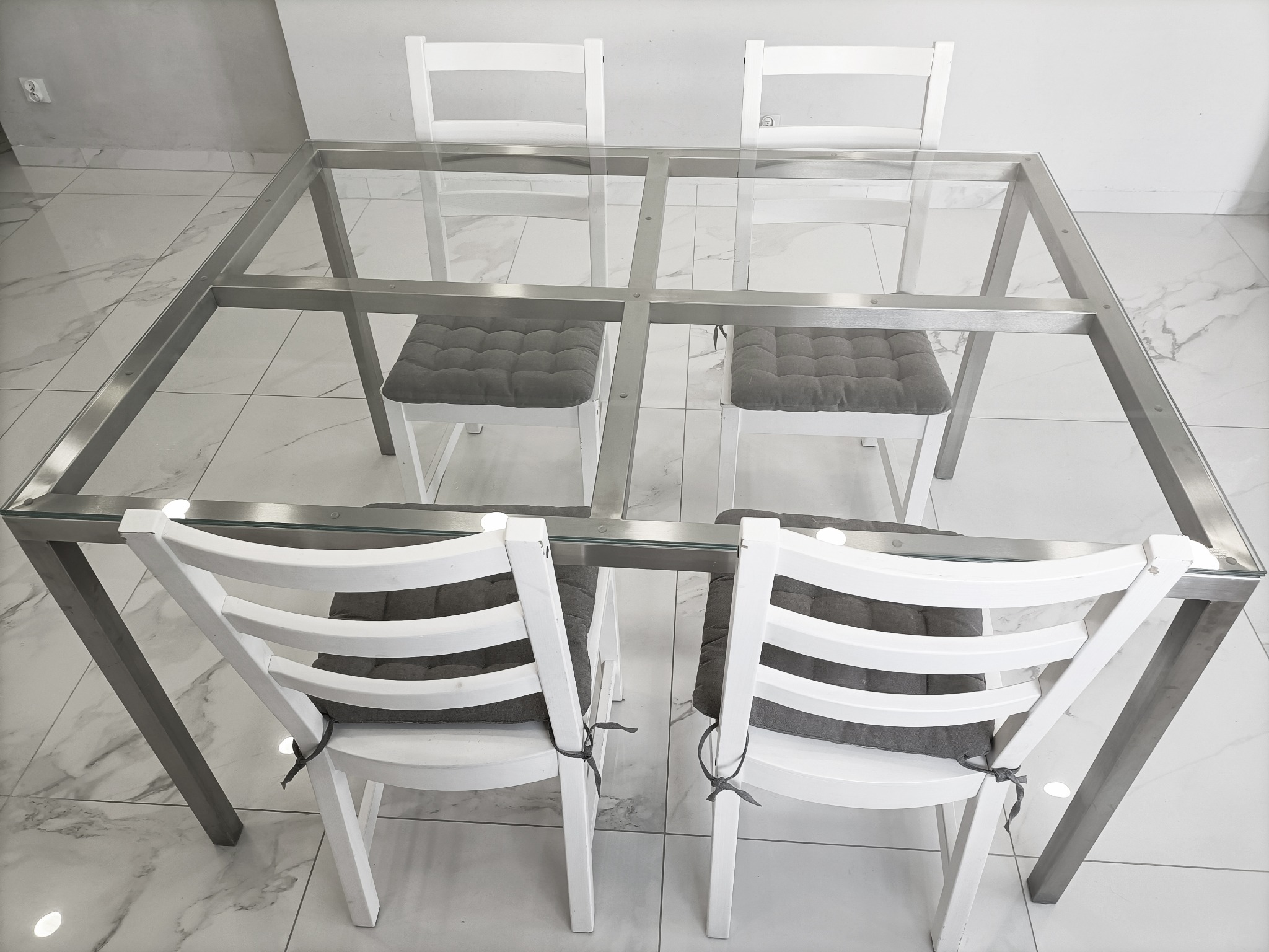 Kwadratowy stół z szklanym blatem i metalową ramą, otoczony czterema białymi krzesłami z szarymi poduszkami, widok z góry na marmurową podłogę.