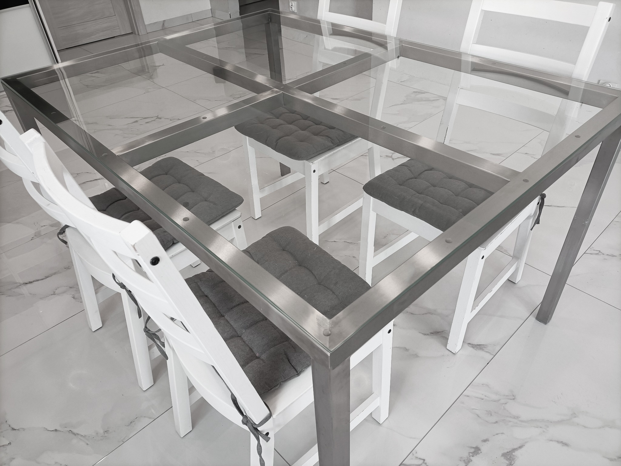 Stół jadalny z przezroczystym szklanym blatem i metalową ramą, otoczony białymi krzesłami z szarymi poduszkami, na tle marmurowej podłogi.