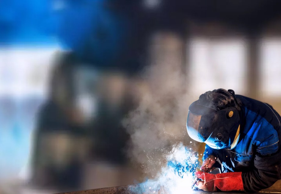 Spawacz w niebieskim kombinezonie i masce spawalniczej, pochylony nad metalową konstrukcją, z iskrami i dymem w powietrzu.