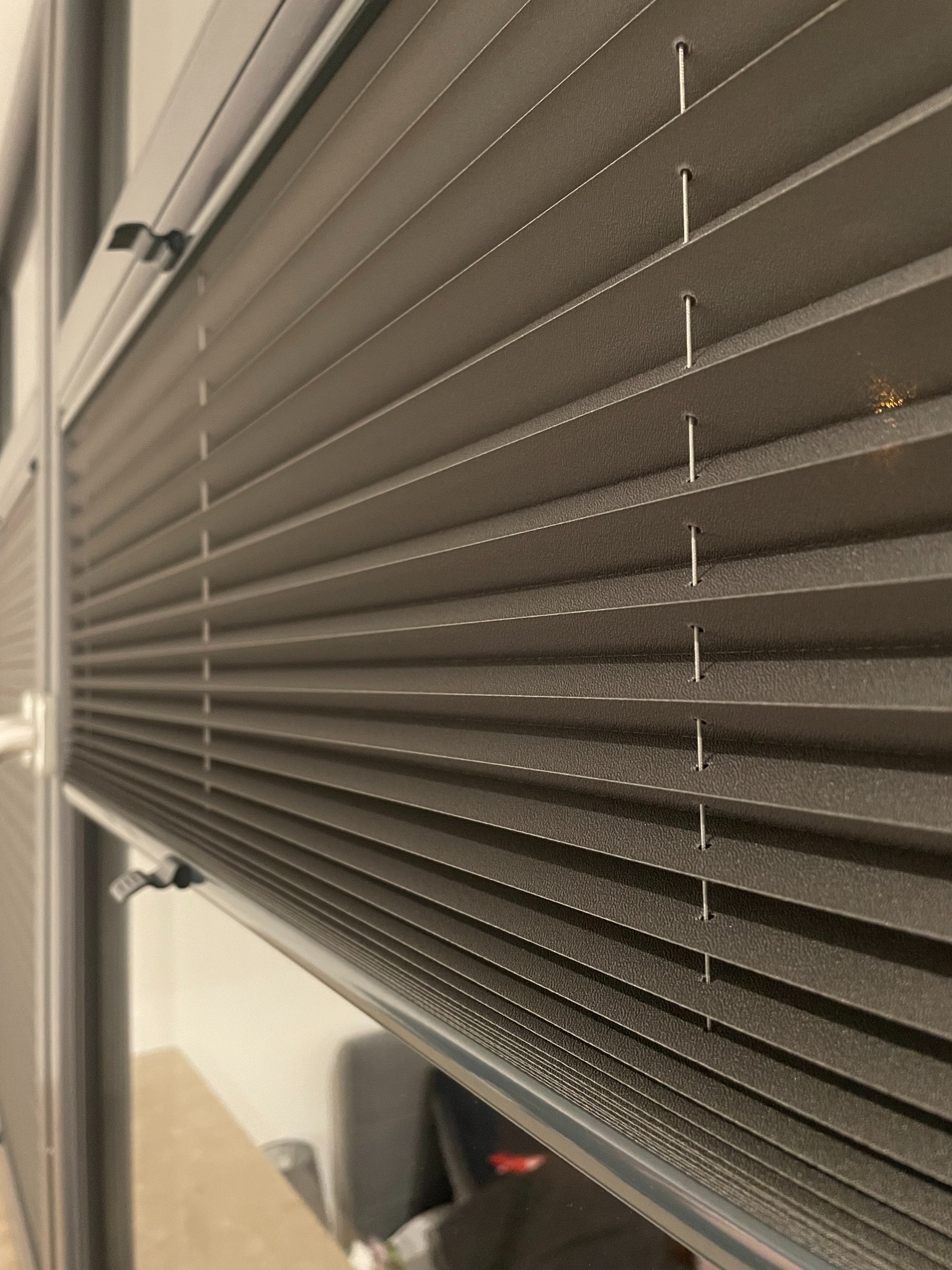 Szczegółowe ujęcie szarej, plisowanej osłony okiennej z widocznymi linkami prowadzącymi przez otwory w materiale, zamontowanej w oknie z szarą ramą.