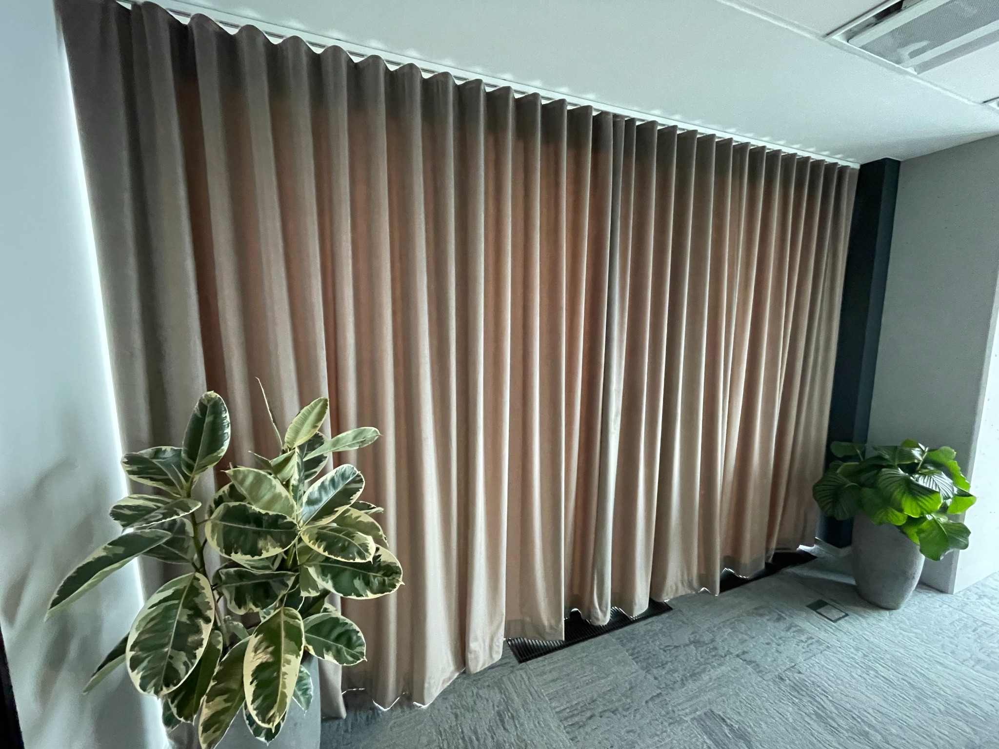 Beżowe, faliste zasłony zakrywające całą ścianę, z widocznymi grzejnikami u dołu, oraz dwoma doniczkami z roślinami po bokach na tle szarej podłogi.