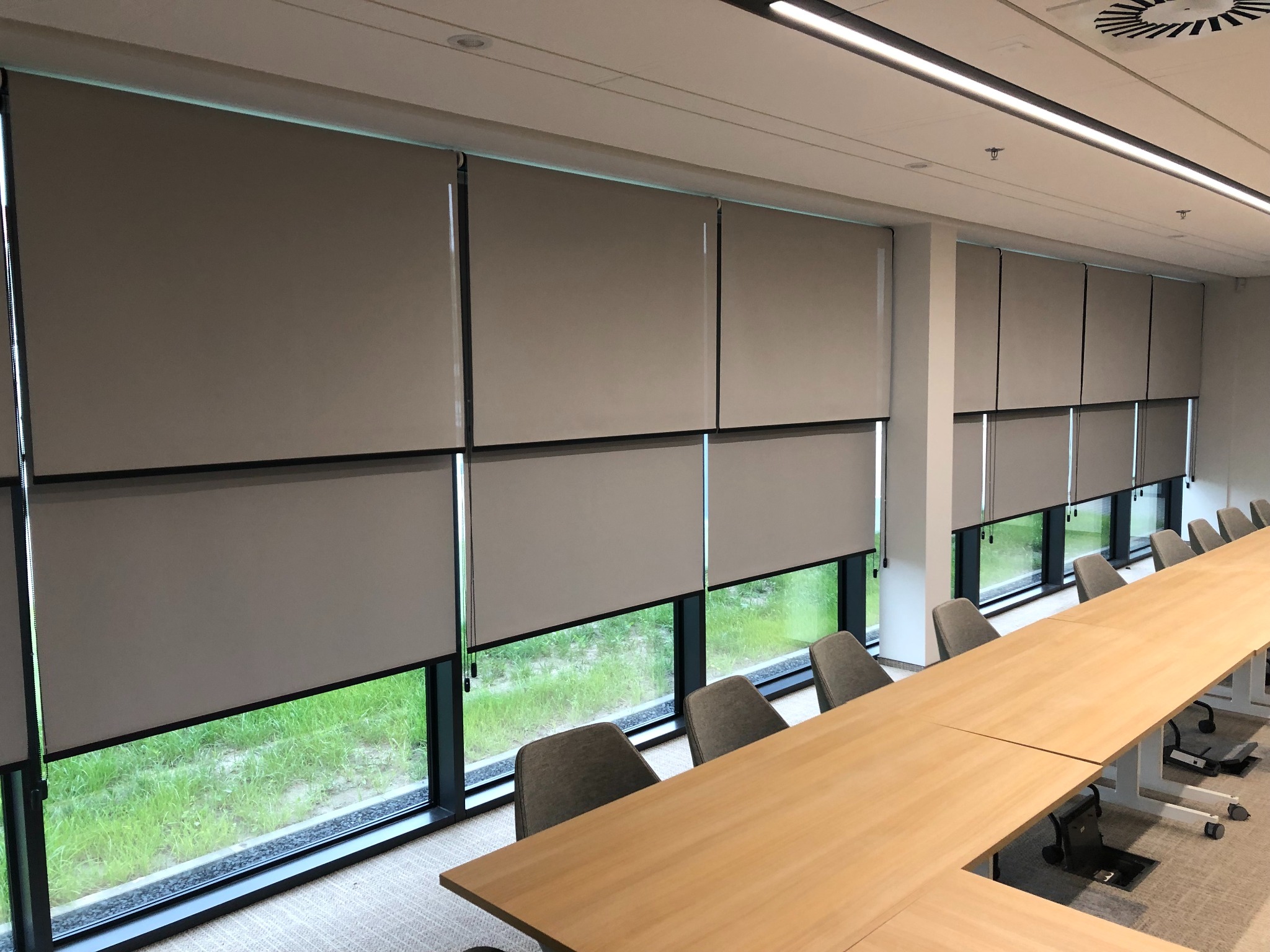 Beżowe rolety materiałowe w kasecie, zamontowane w oknach sali konferencyjnej, częściowo zasłonięte, widok na zieleń za oknem.