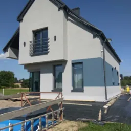 Nowoczesny dom jednorodzinny z szarą elewacją i niebieskim pasem, widoczne przygotowania do zagospodarowania terenu wokół budynku, w tle drzewa i błękitne niebo.