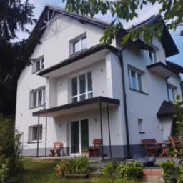 Dwupiętrowy dom z białymi ścianami i ciemnymi elementami wykończeniowymi, widoczny z poziomu ogrodu z drewnianymi meblami.