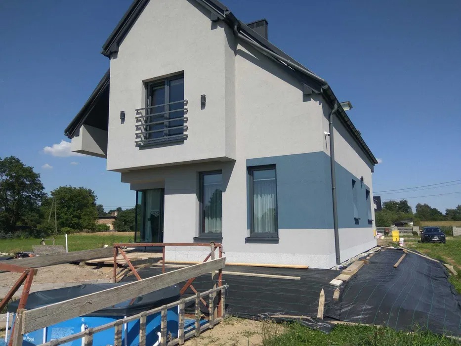 Nowoczesny dom jednorodzinny z szarą elewacją i niebieskim pasem, widoczne przygotowania do zagospodarowania terenu wokół budynku, w tle drzewa i błękitne niebo.