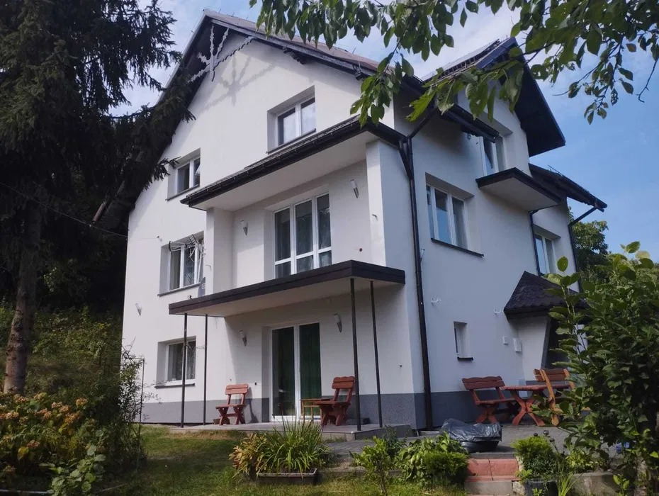 Dwupiętrowy dom z białymi ścianami i ciemnymi elementami wykończeniowymi, widoczny z poziomu ogrodu z drewnianymi meblami.