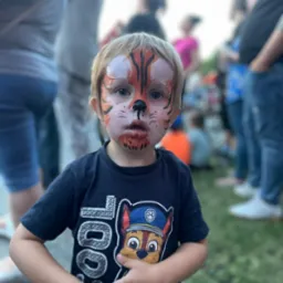 Chłopiec z pomalowaną twarzą w motywy tygrysa, ubrany w ciemną koszulkę z nadrukiem bajkowym, na tle rozmytych postaci.