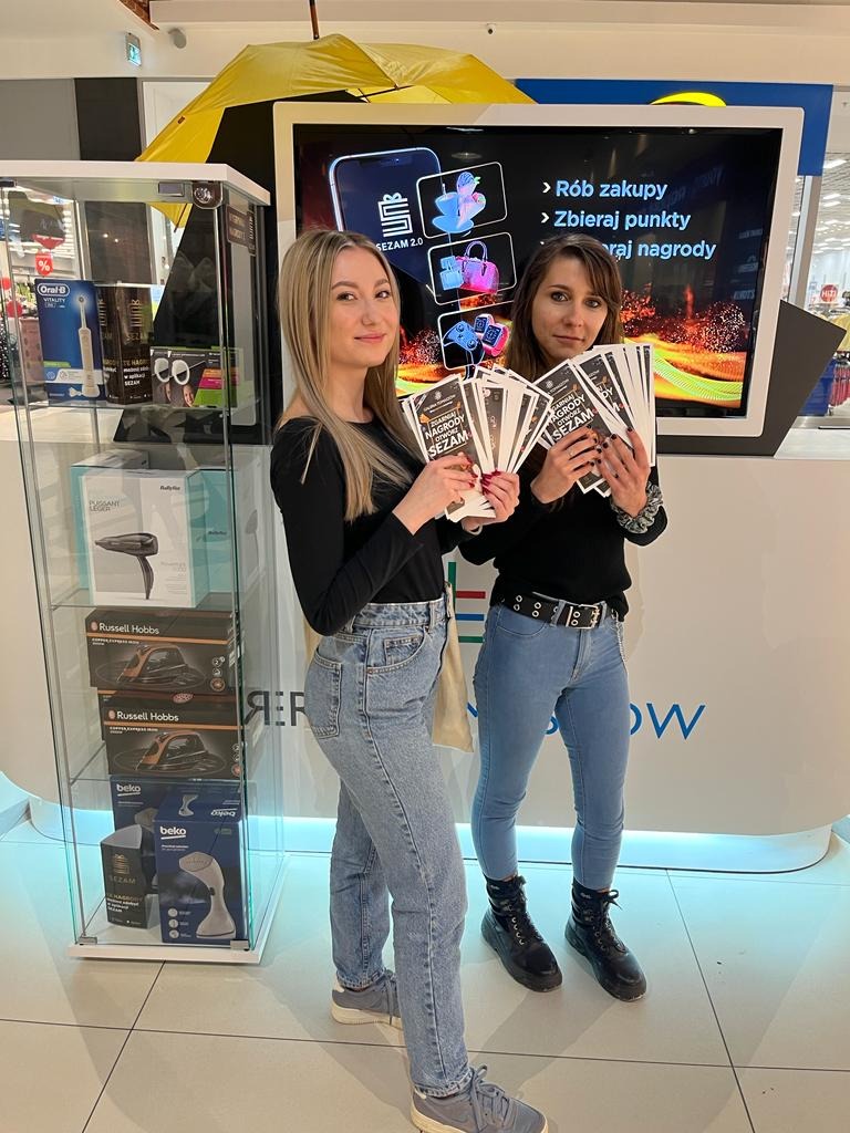 Dwie kobiety prezentują ulotki z napisem 'Nagrody Dziś Sezam' na tle monitora z aplikacją Sezam 2.0 w sklepie z artykułami gospodarstwa domowego i kosmetykami.