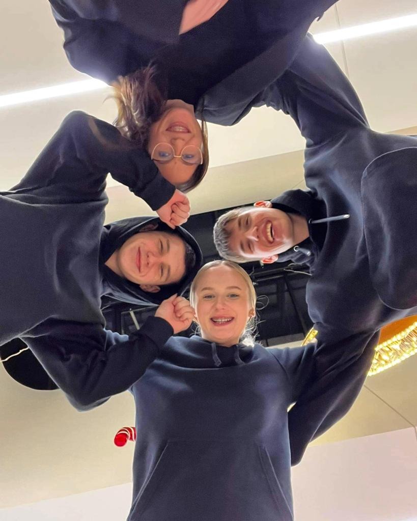 Grupa czterech uśmiechniętych młodych osób w ciemnych bluzach z kapturem, stojących w kręgu i patrzących w dół w obiektyw, z widocznym fragmentem dekoracji świątecznej w tle.