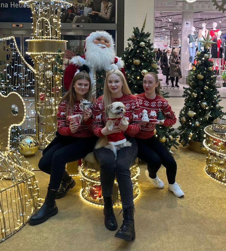 Trzy kobiety w świątecznych swetrach z reniferami pozują z psem ubranym w czerwony sweterek, obok dekoracji świątecznych i figury Świętego Mikołaja w centrum handlowym.