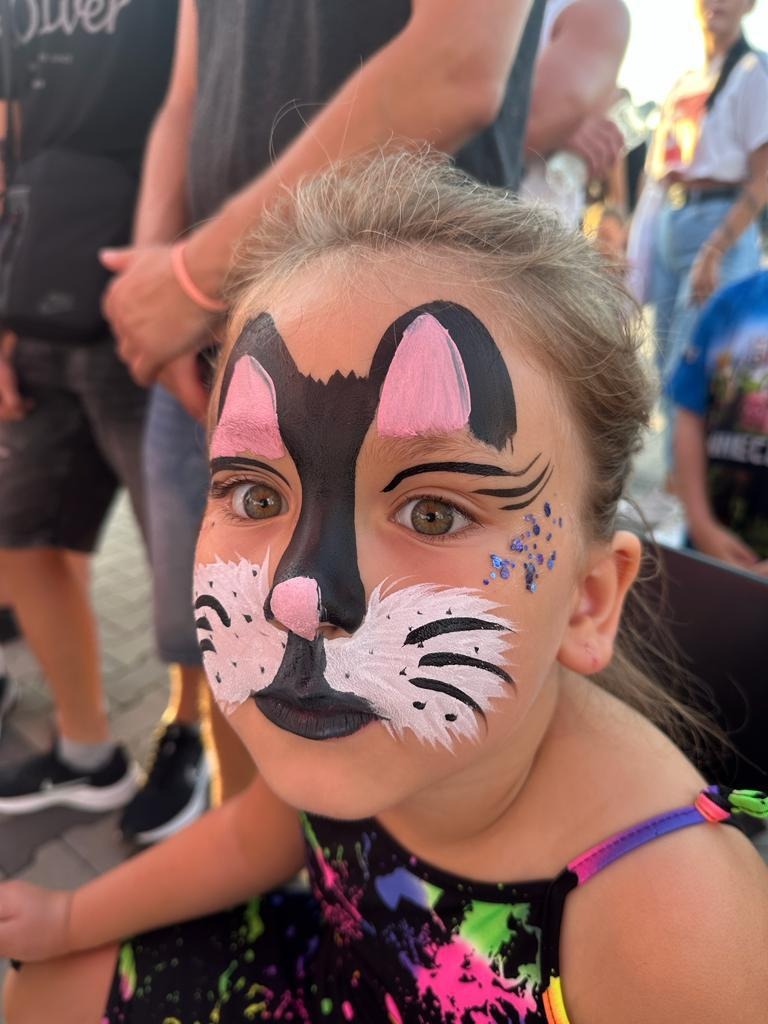 Dziewczynka z pomalowaną twarzą w motywy kota, z czarnym nosem, różowymi uszami, białymi wąsami i brokatem pod okiem.