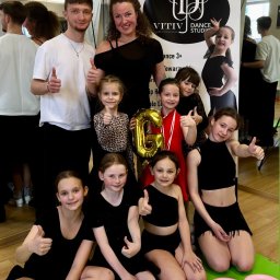 Vitiv Dance Studio - Uśmiechnięta grupa dzieci i dwoje dorosłych w strojach tanecznych, pokazujących kciuki w górę, na tle lustrzanej ściany i baneru 'VITIV DANCE STUDIO'. Na zielonej macie tort urodzinowy i talerzyki,...