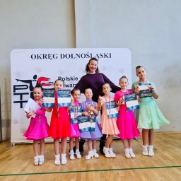 Vitiv Dance Studio - Uśmiechnięta trenerka z sześcioma młodymi tancerkami w kolorowych sukienkach, dumnie prezentującymi dyplomy i medale po turnieju tańca na tle baneru Polskiego Towarzystwa Tanecznego Okręgu...