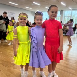 Vitiv Dance Studio - Trzy uśmiechnięte dziewczynki w jaskrawych strojach tanecznych (neonowy żółty, lawendowy, czerwony) pozują na drewnianym parkiecie, z rozmazanym tłem innych dzieci i dorosłych.