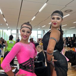 Vitiv Dance Studio - Dwie tancerki w błyszczących strojach turniejowych: młodsza w różowej sukience z kryształkami, starsza w czarnej z frędzlami, pozują pewnie na tle sali tanecznej.