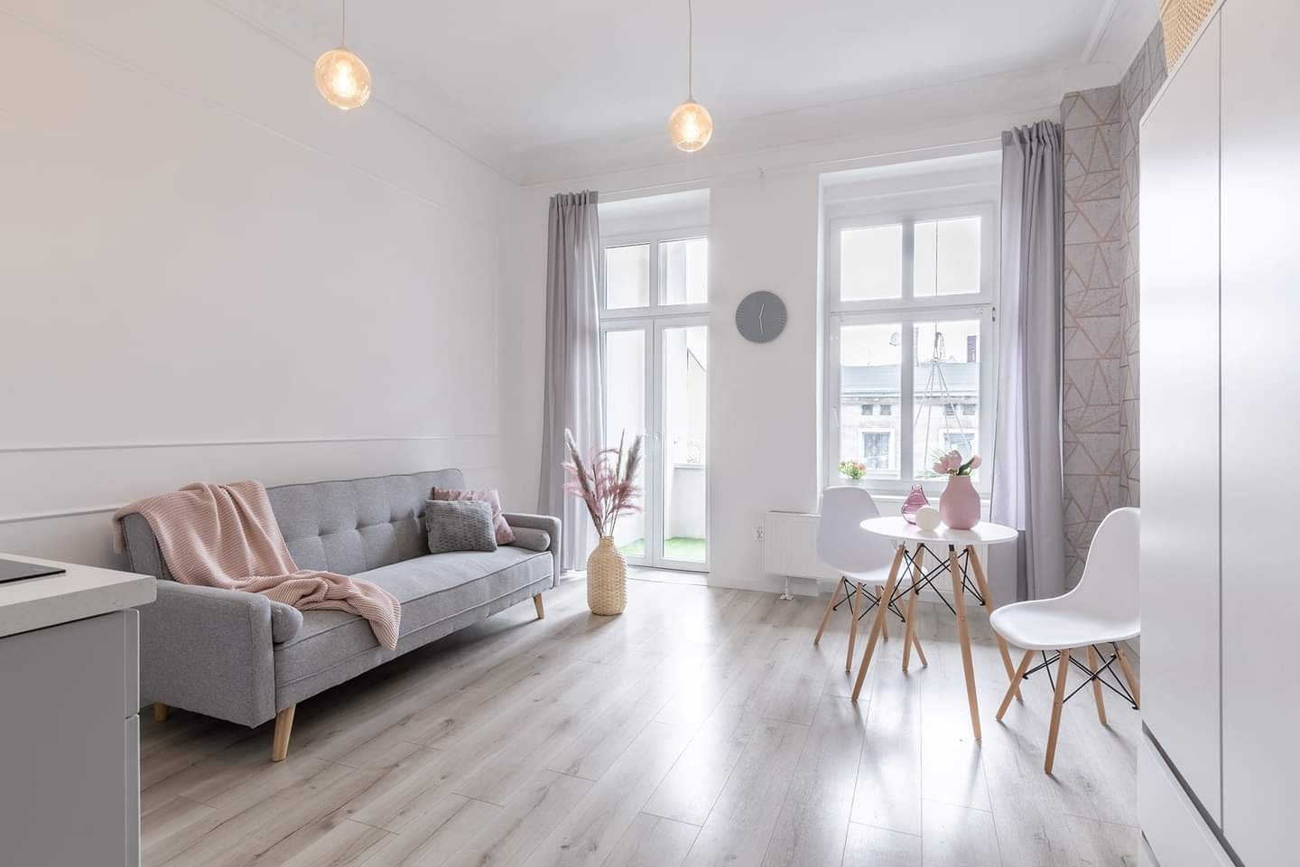 Jasne, przestronne wnętrze po remoncie: szara sofa z różowym pledem, okrągły stolik z kwiatami, drewniana podłoga, białe ściany z dekoracyjną listwą, lampy wiszące i geometryczna tapeta