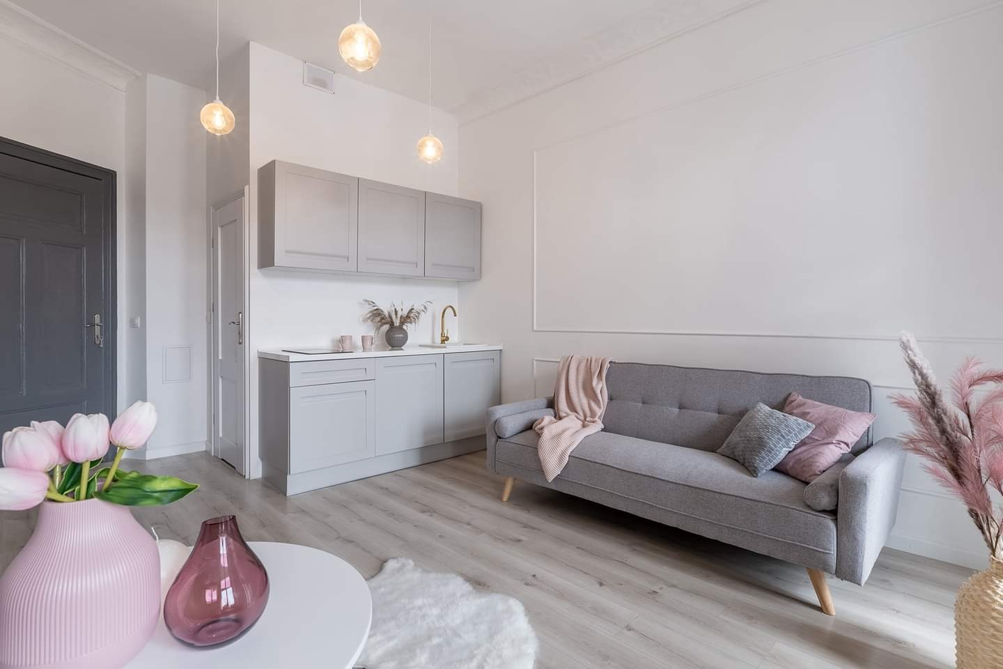Jasne wnętrze po remoncie z szarą zabudową kuchenną, szarą sofą i kwiatami w wazonach w odcieniach różu. Widoczne trzy wiszące lampy kuliste i panele podłogowe.