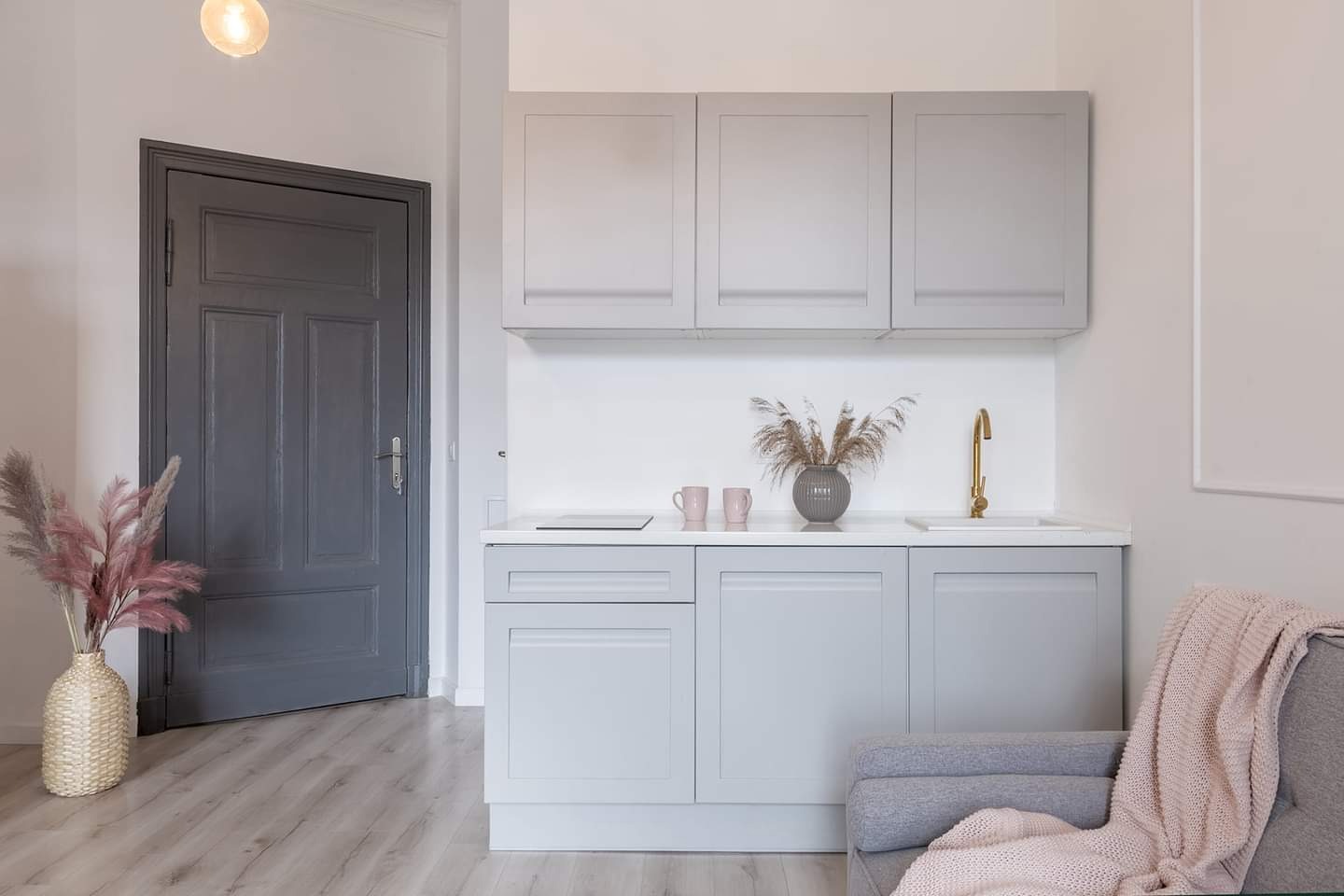 Jasnoszara, minimalistyczna kuchnia z drewnianą podłogą, złotą baterią i bukietem suszonych traw w wazonie na blacie, obok szarych szafek i ciemnoszarych drzwi.