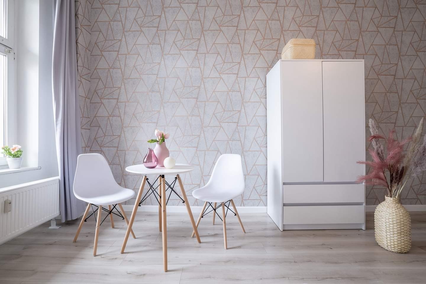 Jasne wnętrze pokoju z geometryczną tapetą, białymi krzesłami i okrągłym stolikiem z różowymi wazonami, obok biała szafa i dekoracyjny wazon z suszonymi kwiatami.
