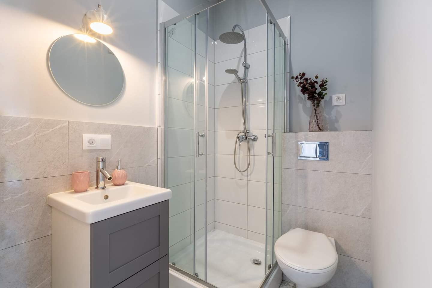 Jasna łazienka po remoncie: szklana kabina prysznicowa z deszczownicą, umywalka z szarą szafką, okrągłe lustro z oświetleniem, płytki imitujące marmur, wisząca toaleta.