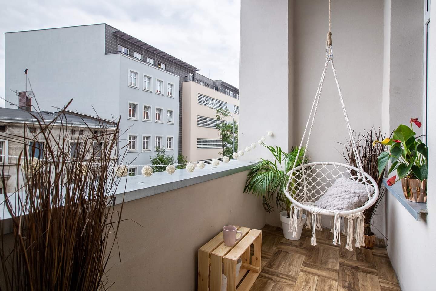 Przytulny balkon z drewnianą podłogą, wiszącym fotelem hamakowym, roślinami w doniczkach i dekoracyjnymi kulami świetlnymi, widok na budynki mieszkalne