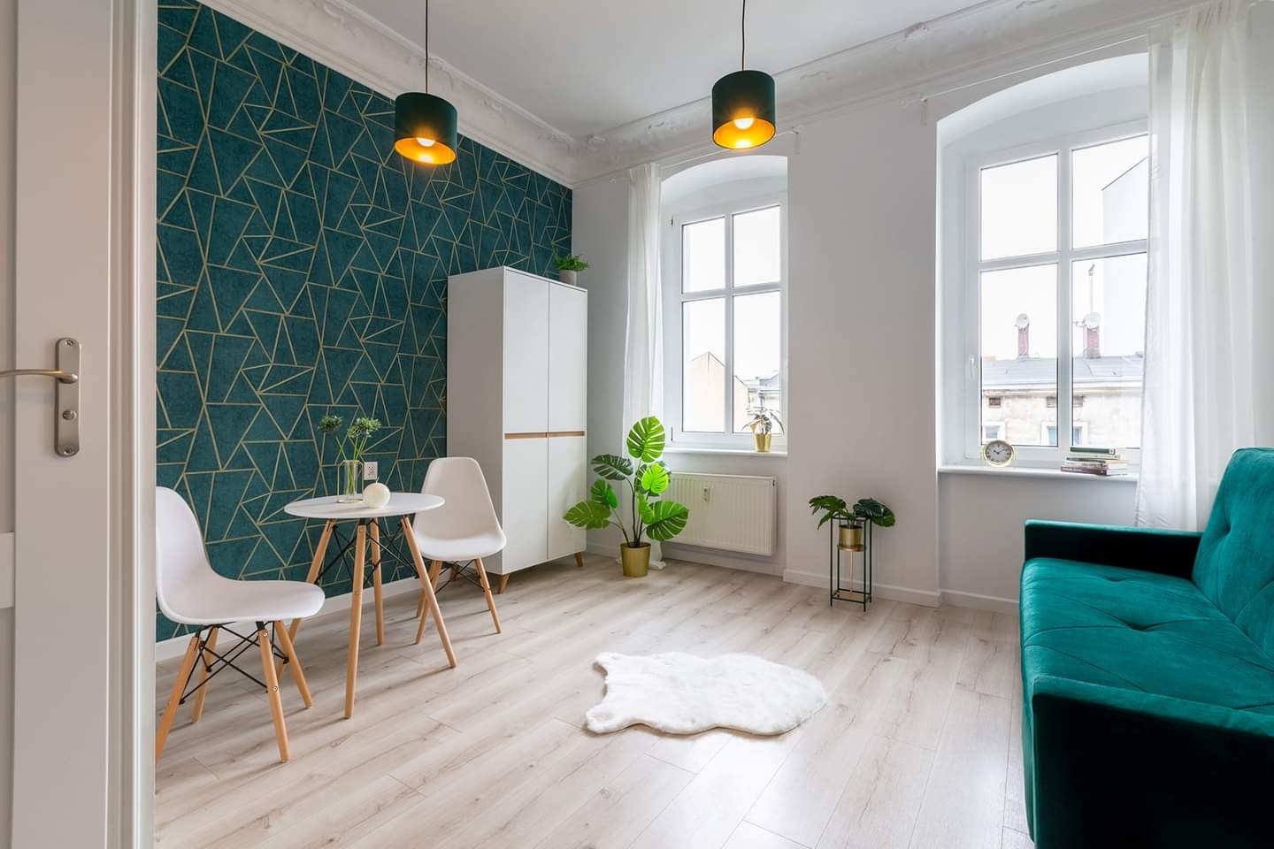Jasne wnętrze pokoju z turkusową ścianą w geometryczne złote wzory, białymi meblami, drewnianą podłogą i zieloną kanapą, oświetlone wiszącymi lampami z zielonymi abażurami.