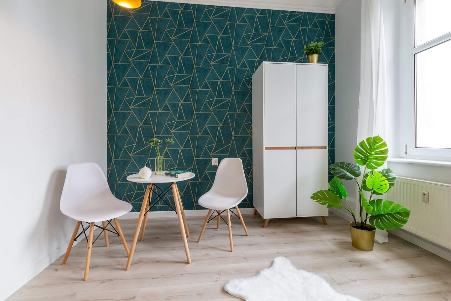Nowoczesny pokój z turkusową ścianą w geometryczne złote wzory, białymi krzesłami i stolikiem, białym regałem z drewnianymi akcentami oraz roślinami doniczkowymi, oświetlony ciepłym światłem.