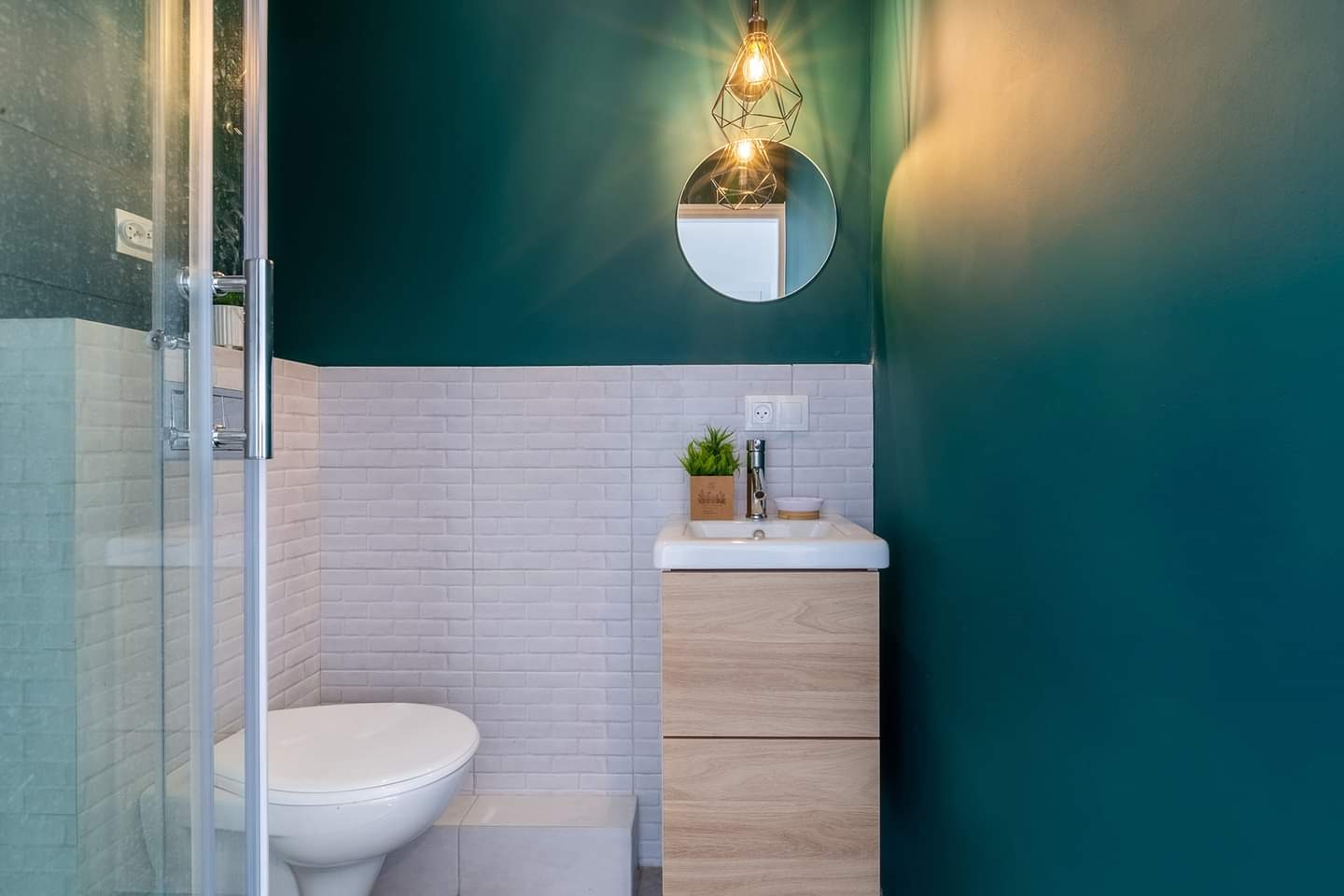 Nowoczesna łazienka z turkusową ścianą, białymi cegiełkowymi płytkami, umywalką z szafką z jasnego drewna i geometryczną lampą wiszącą