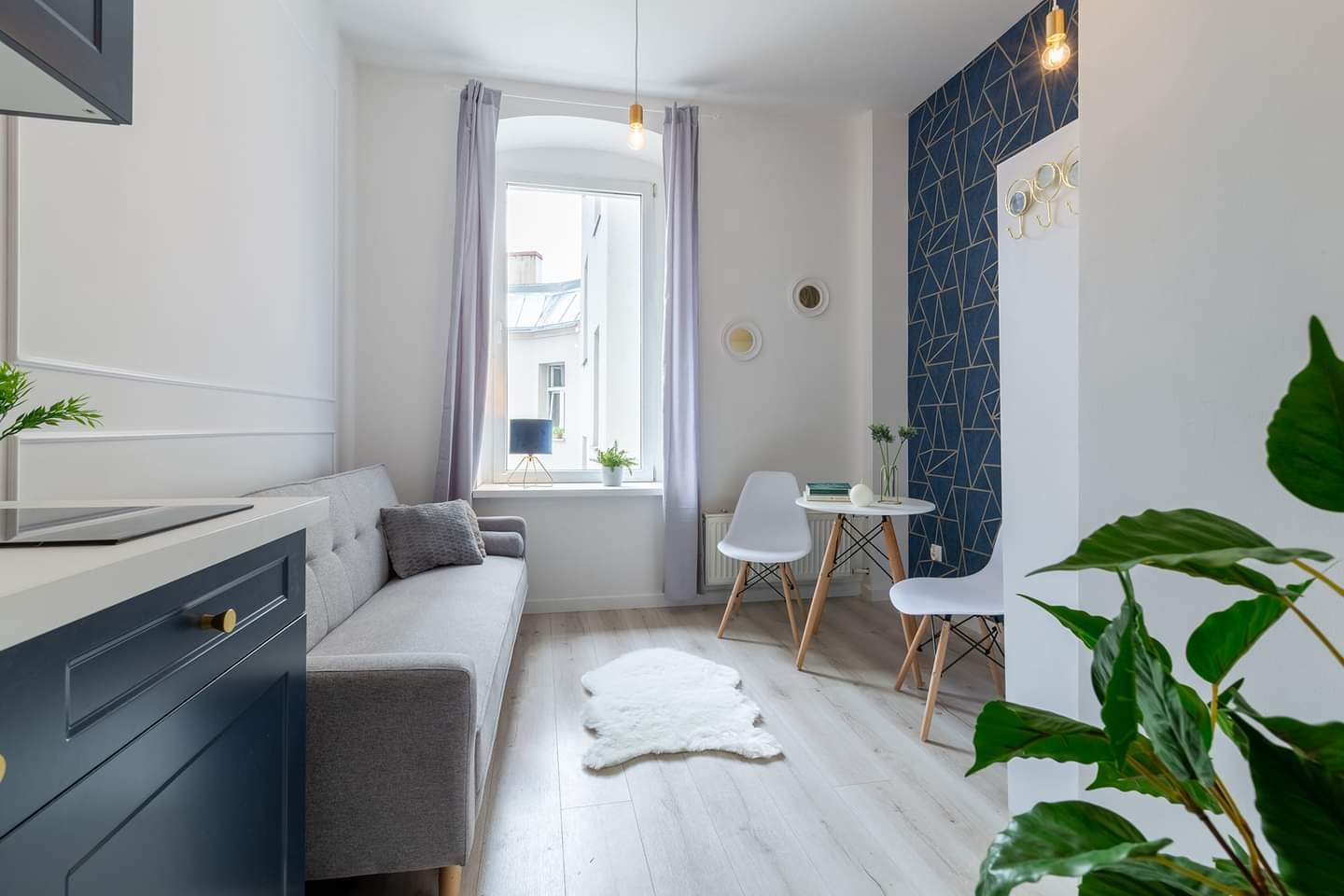 Jasne, nowocześnie urządzone wnętrze kawalerki z szarą sofą, białym stolikiem i ścianą z geometryczną tapetą w odcieniach granatu i złota.
