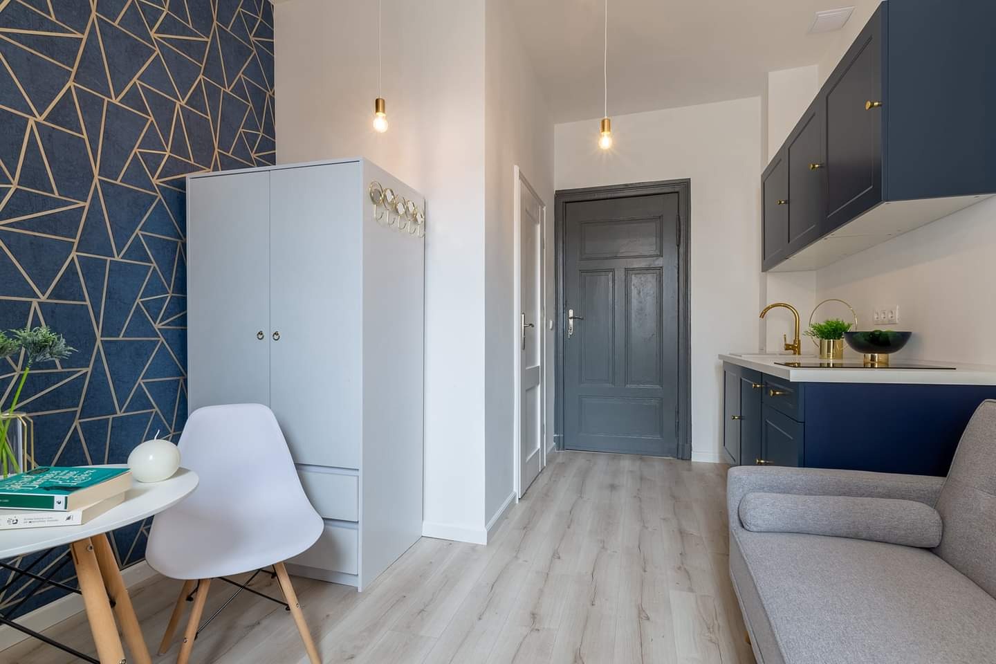 Nowoczesne wnętrze kawalerki po remoncie: granatowa ściana z geometrycznym wzorem, jasnoszara szafa z wieszakami, białe krzesło przy okrągłym stoliku, aneks kuchenny z granatowymi szafkami i złotą...