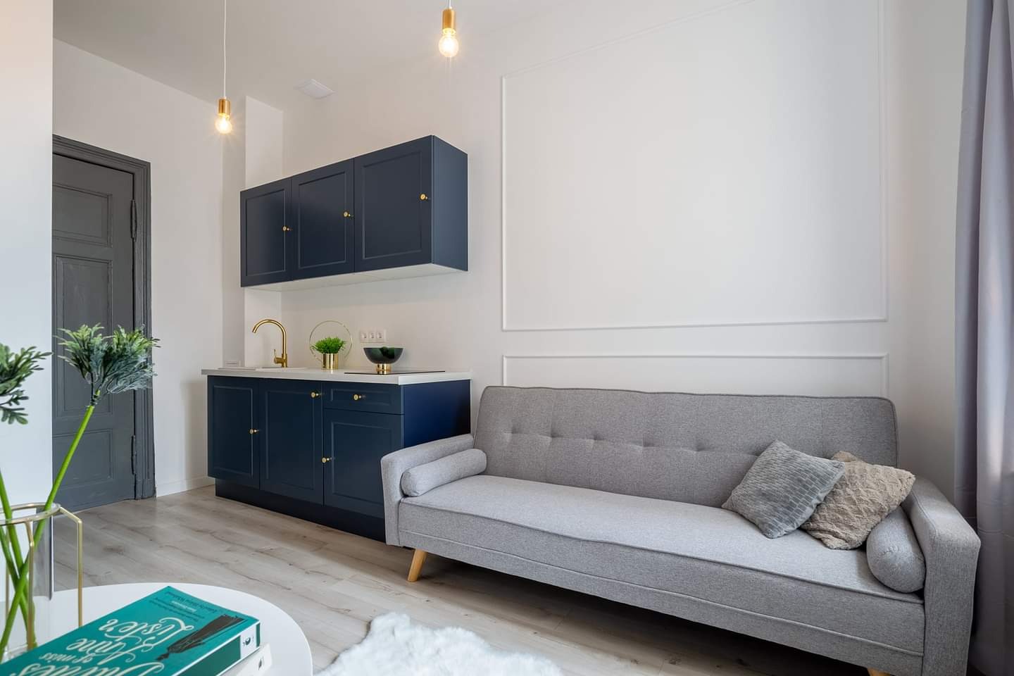 Nowoczesne wnętrze kawalerki z aneksem kuchennym w kolorze granatowym, szarą sofą i białymi ścianami z dekoracyjnymi listwami.