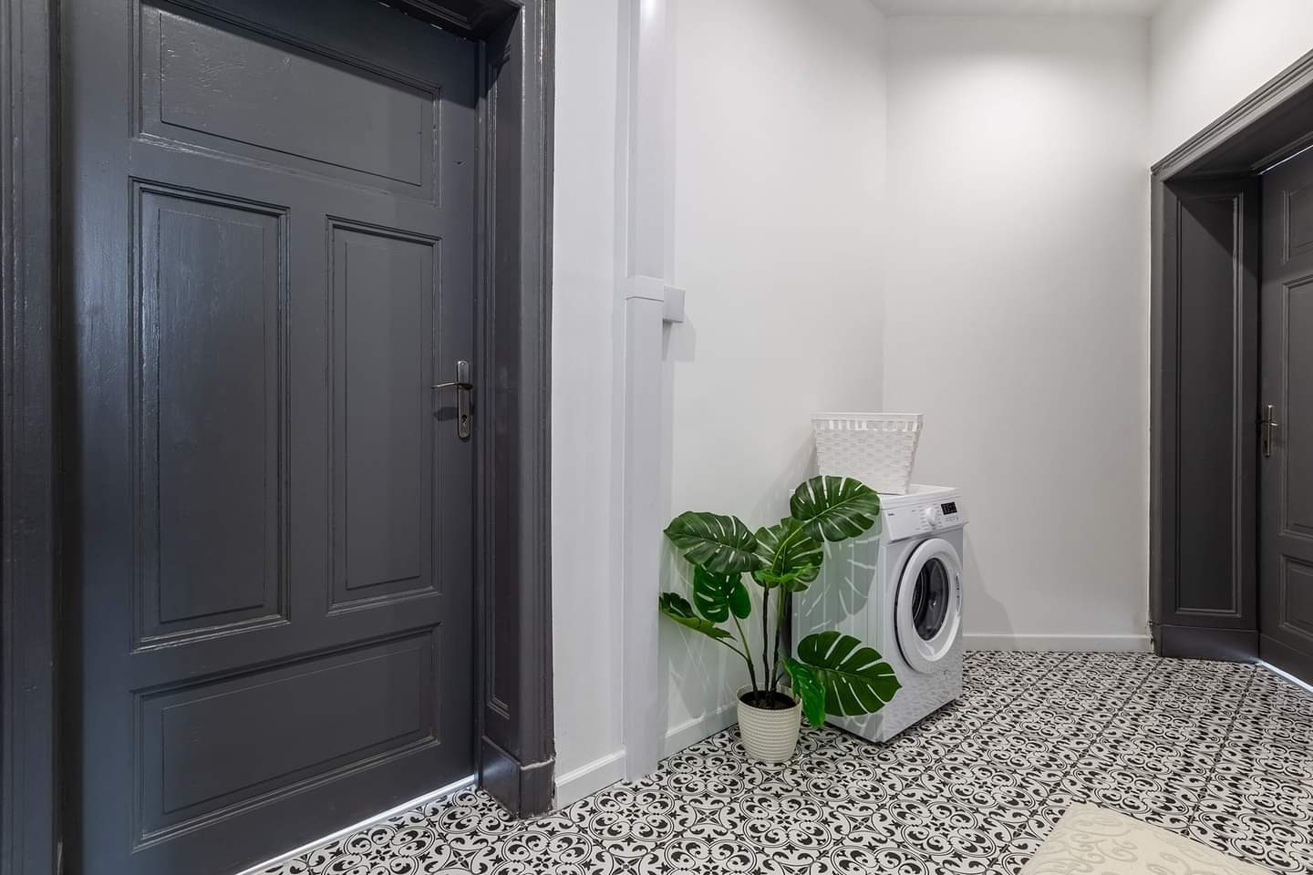 Wnętrze przedpokoju z odnowionymi, ciemnoszarymi drzwiami, wzorzystą czarno-białą podłogą, białą ścianą z pralką i rośliną doniczkową.