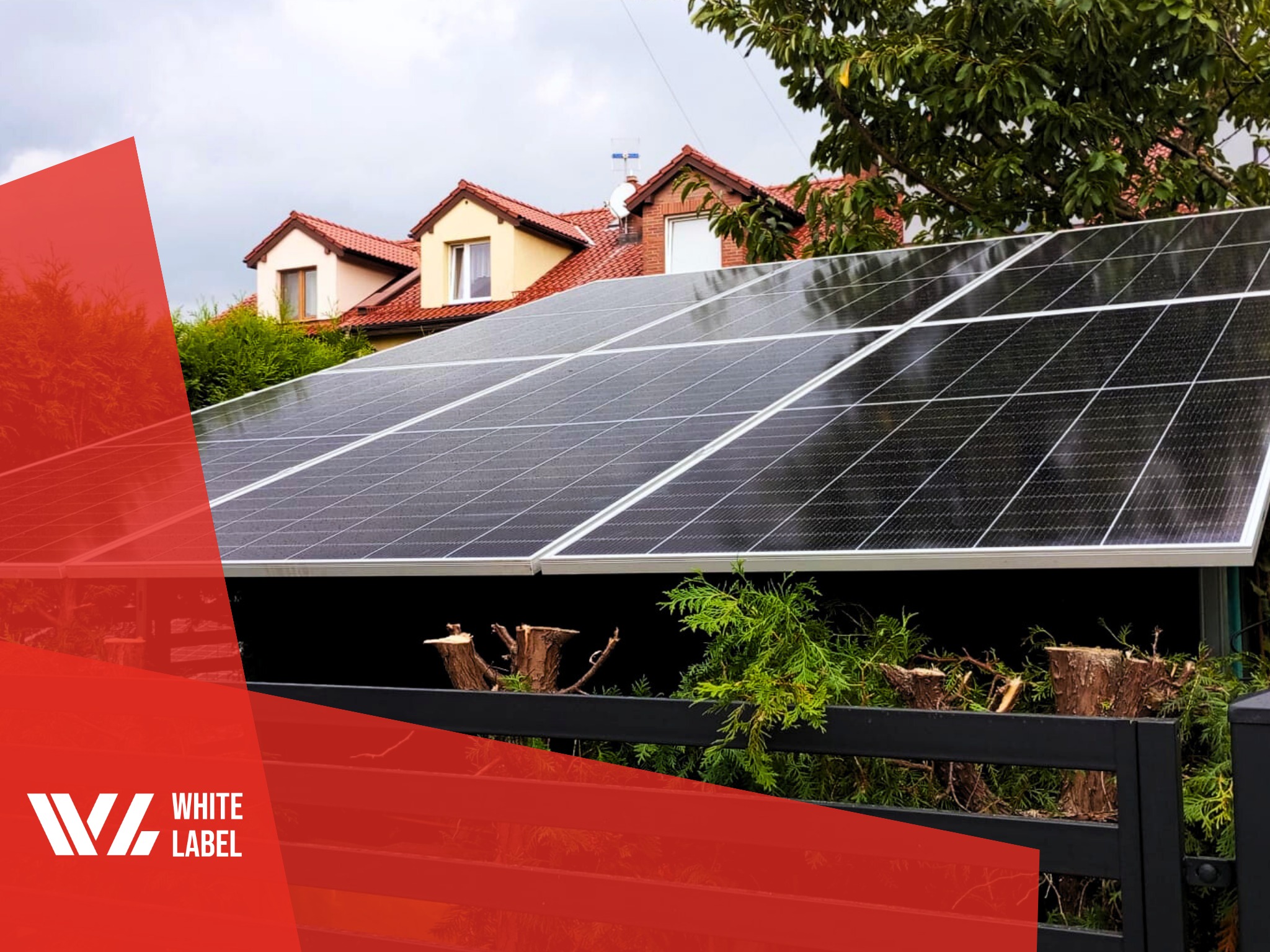 Panele słoneczne zamontowane na dachu domu jednorodzinnego z czerwonymi dachówkami, widoczne przez czarne ogrodzenie z przyciętymi krzewami; w lewym dolnym rogu logo firmy.