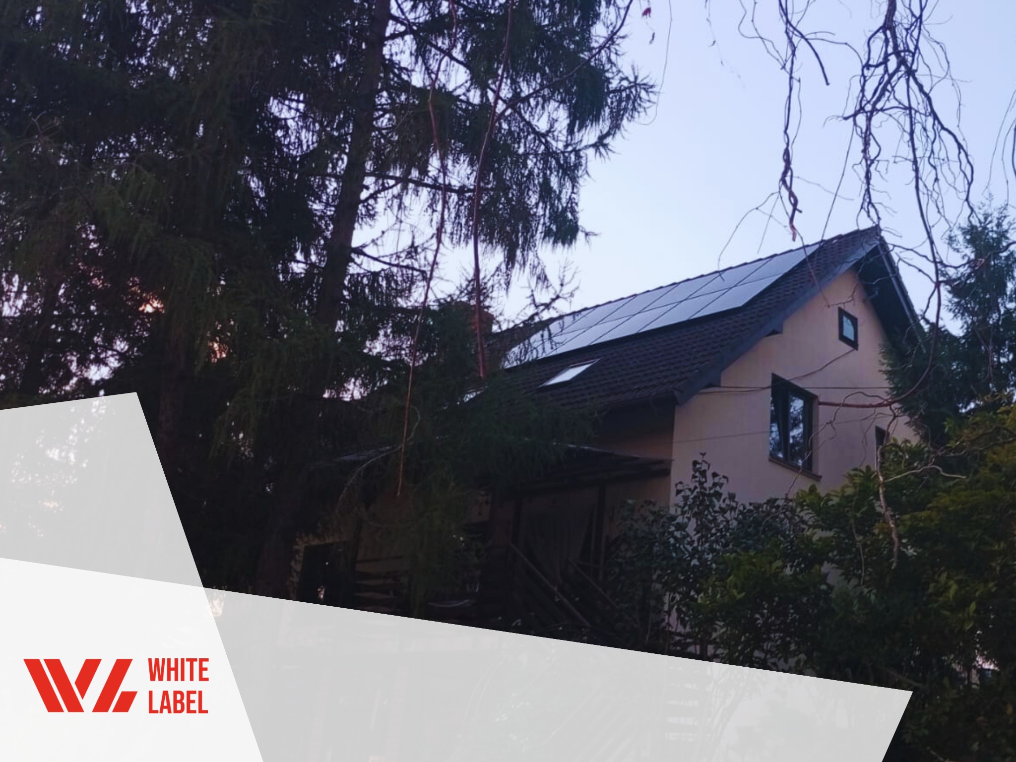 Panele słoneczne na dachu dwuspadowym domu jednorodzinnego, widoczne zza drzew, z logo firmy w lewym dolnym rogu.